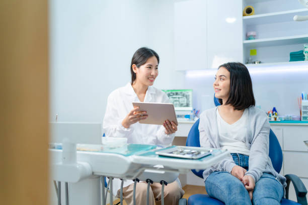 Asian young female dentist holding digital tablet and giving advice or consultation to Caucasian young girl patient. Doctor explaining about oral care treatment service that provide in dental clinic.